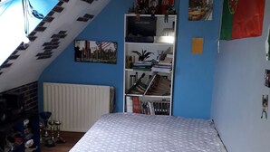 Ein Schlafzimmer mit schrägen Wänden, einem Bett mit gemustertem Überwurf, einem Bücherregal, einem Heizkörper und einem Stoppschild an der Wand.