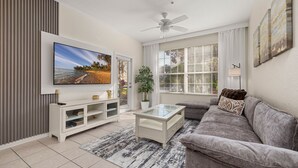 A living room with a flat-screen TV, a ceiling fan, a grey sofa, a coffee table, a TV stand, and a large window with blinds.