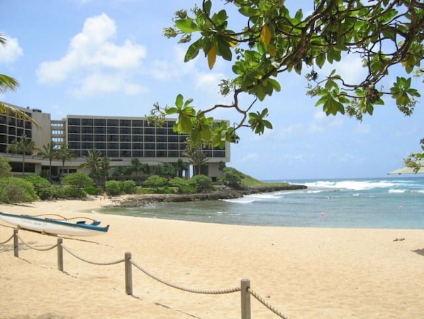 A beach with a building in the background, a rope barrier, and a boat on the sand.
