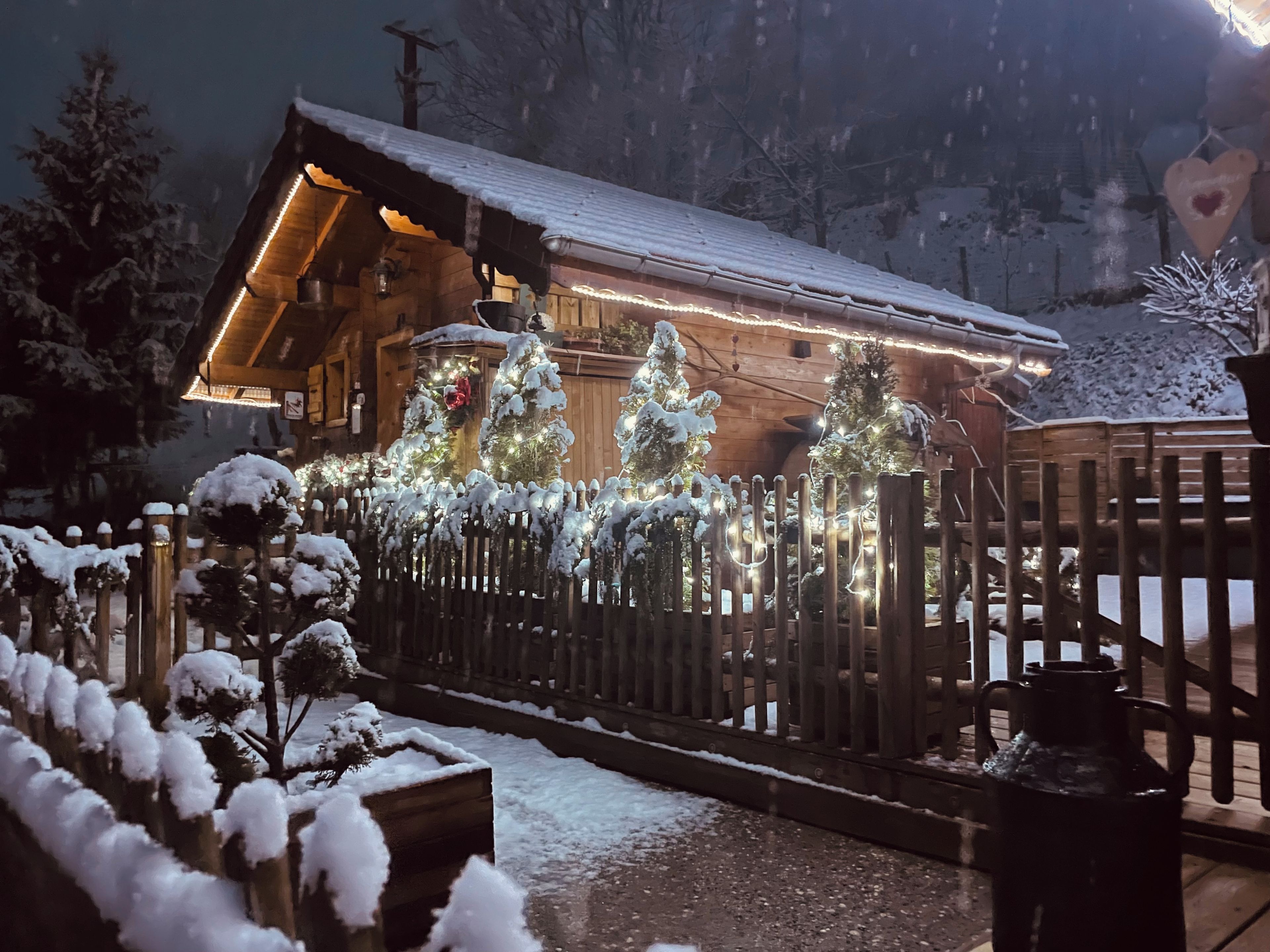 A wooden cabin with a snow-covered roof, surrounded by a wooden fence and snow-covered trees.