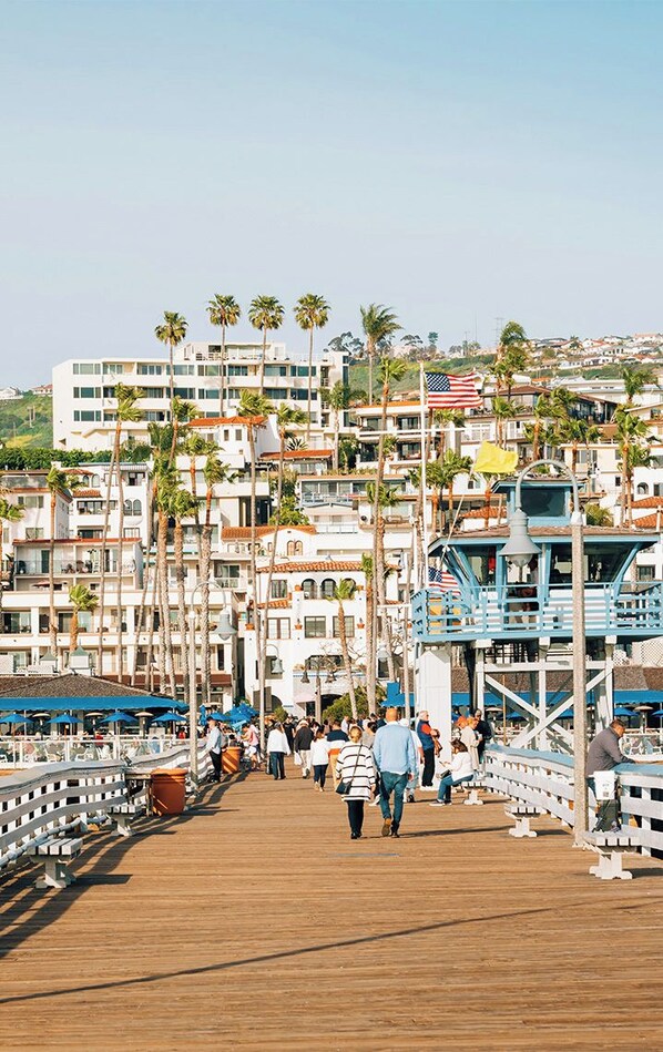 Un paseo marítimo de madera con gente caminando, palmeras y edificios al fondo.