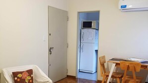 A room with a wooden floor, a white chair with a floral pillow, a table with chairs, a white air conditioner, and a door leading to another room.
