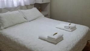 A neatly made bed with white linens and towels, a white wardrobe, and a window with sheer curtains.