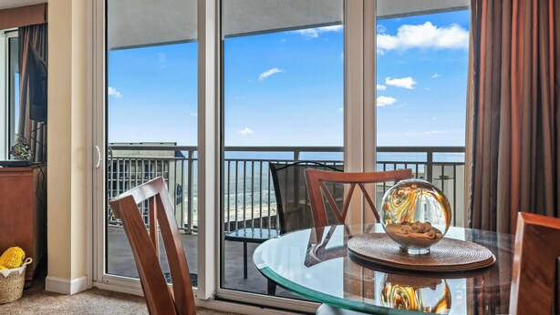A balcony with a glass table and chairs, a bowl of cookies, and a view of the beach.