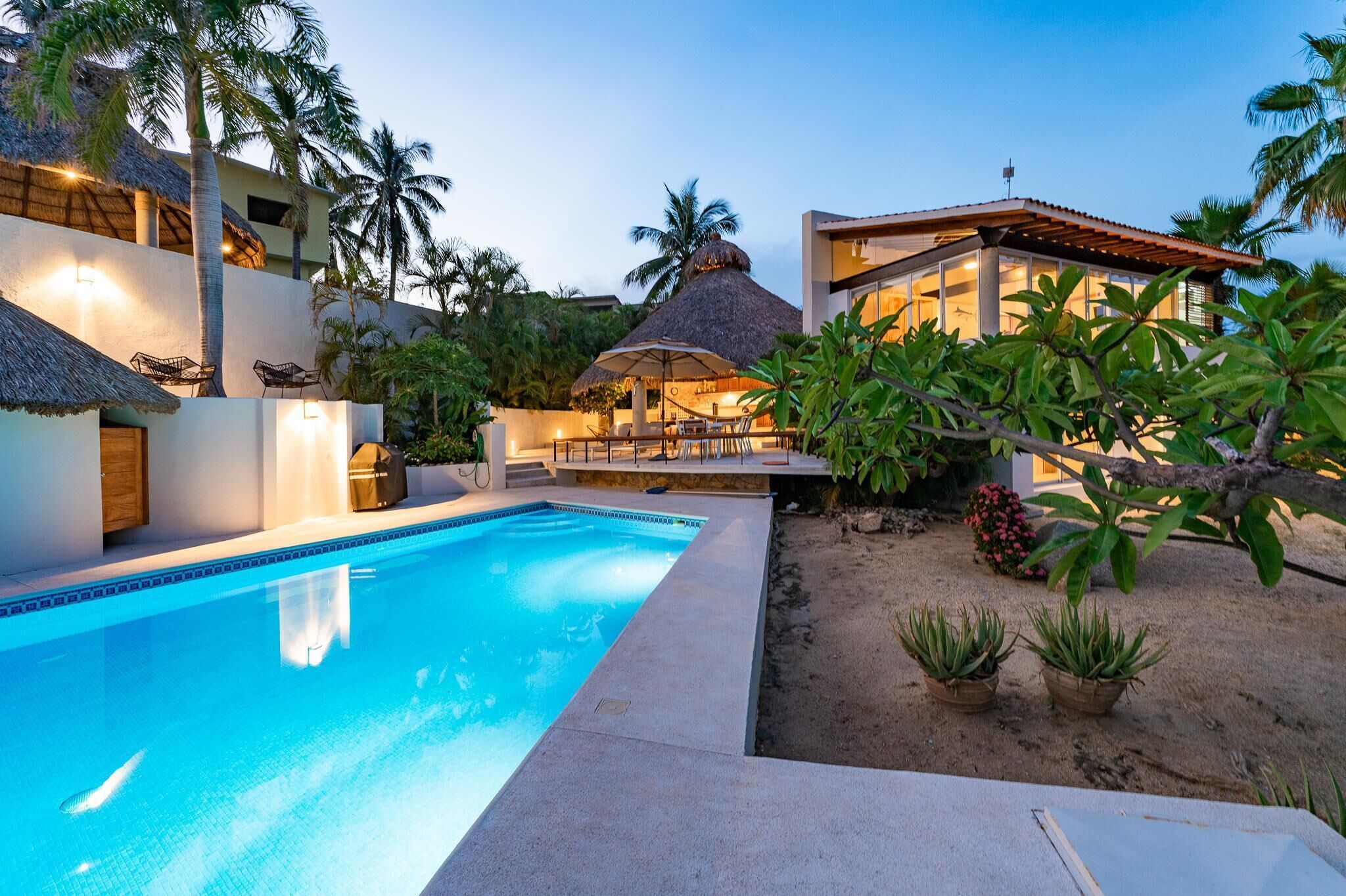 A swimming pool with a thatched roof structure, surrounded by palm trees and potted plants.