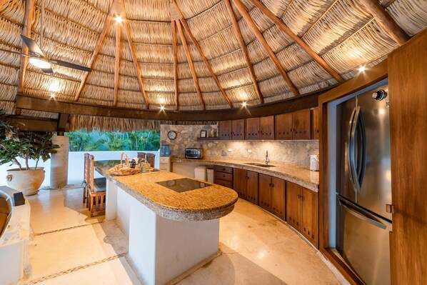 A kitchen with a large island, wooden cabinets, a stainless steel refrigerator, and a thatched roof ceiling.