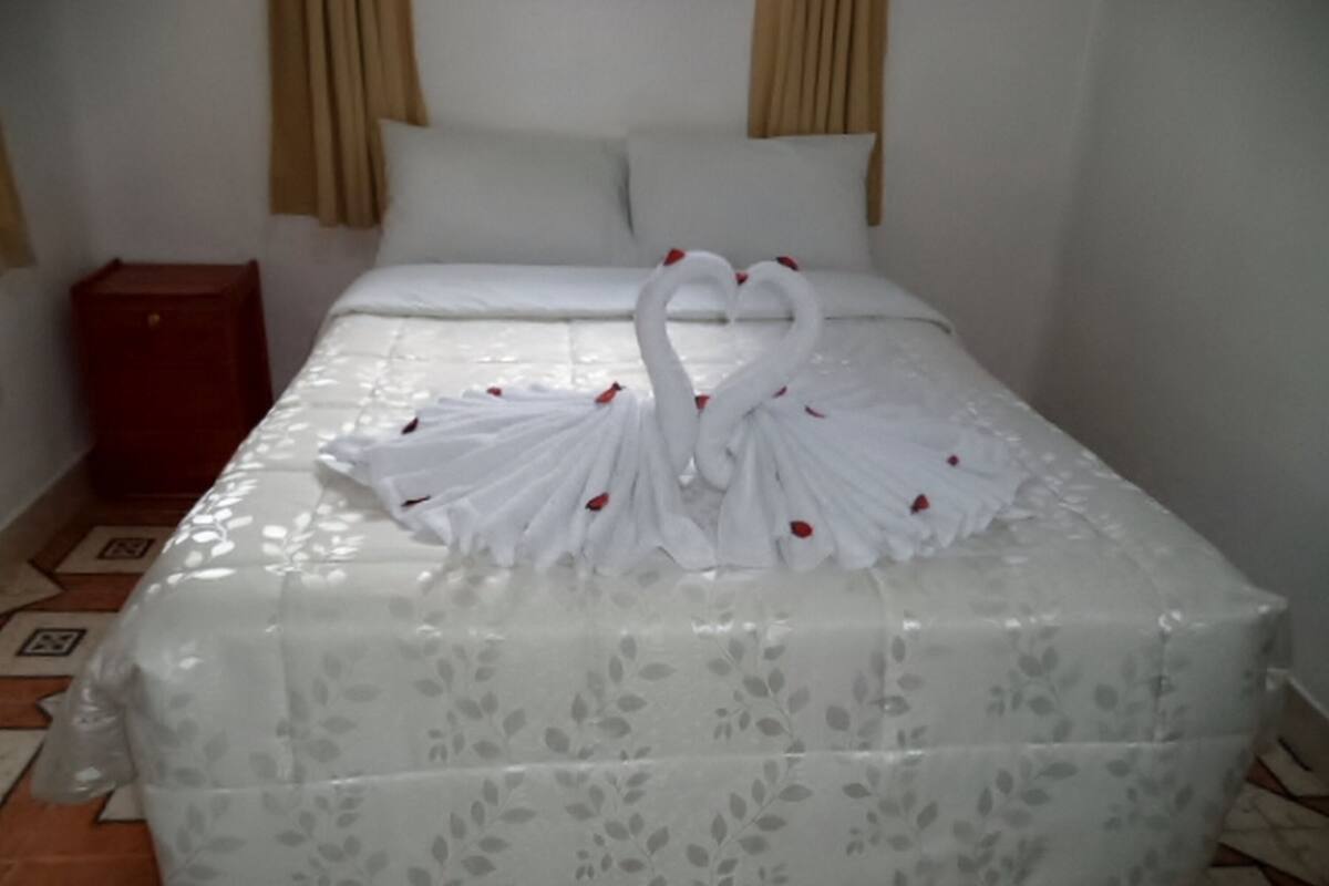 A bed with a white floral bedspread and a towel arrangement in the shape of two swans with red flowers.