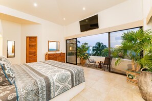 Un dormitorio con cama, cómoda, televisor montado en la pared y balcón con plantas en macetas.