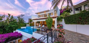 Una casa moderna con piscina, zona de estar exterior y exuberante vegetación.