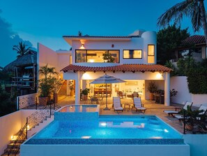 Una casa de dos pisos con piscina, mobiliario exterior y una escalera que conduce al nivel superior.