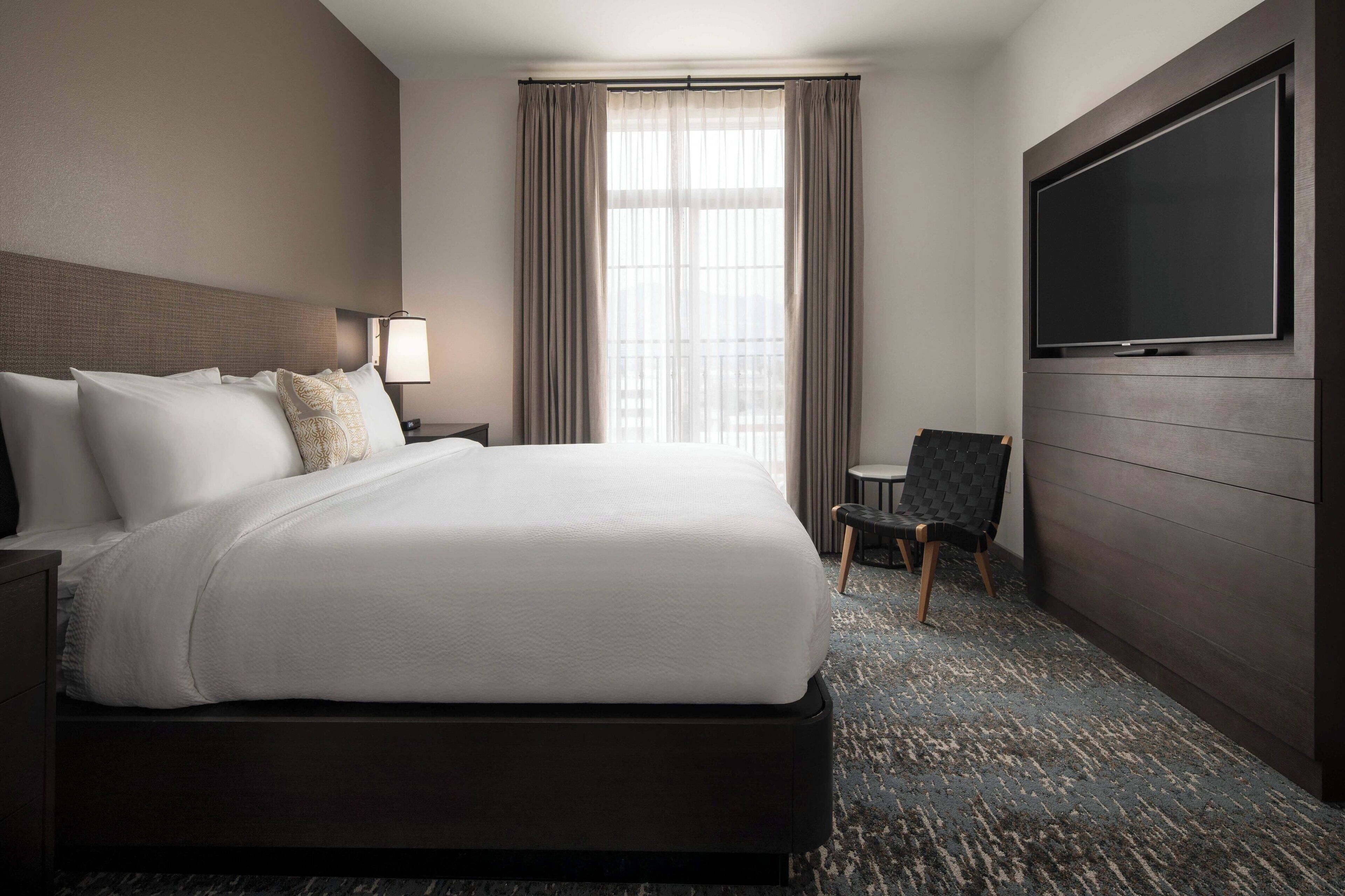 A hotel room with a large bed, a TV mounted on the wall, a chair, and a window with curtains.