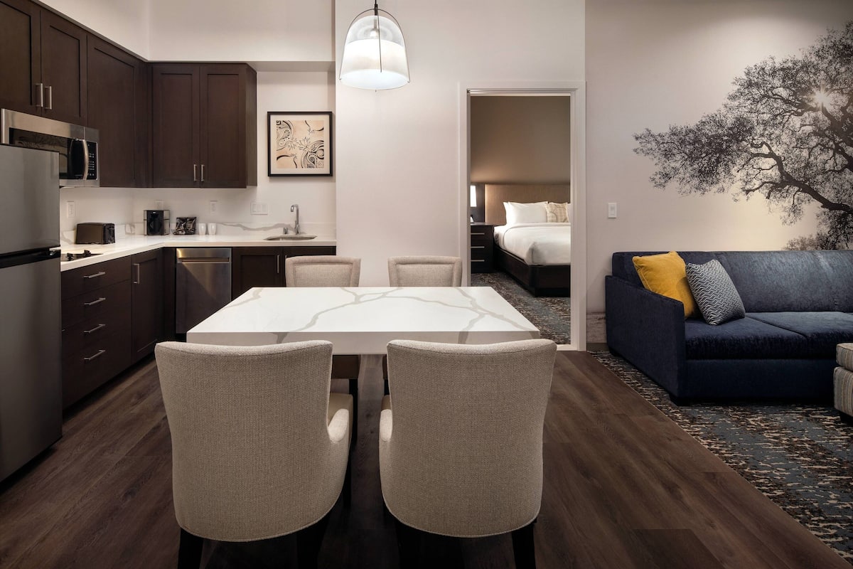 Una cocina moderna con armarios de madera oscura, encimera blanca y comedor con mesa blanca y cuatro sillas. En la sala de estar, hay un sofá azul oscuro con un tapete estampado.