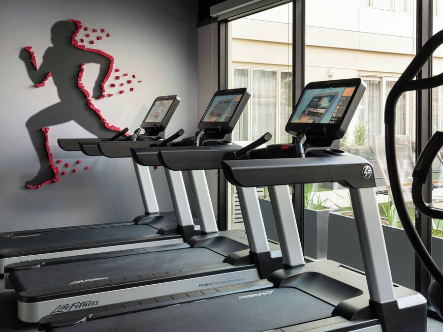 Three treadmills with digital screens, a wall art of a running silhouette, and a window with a view of buildings.