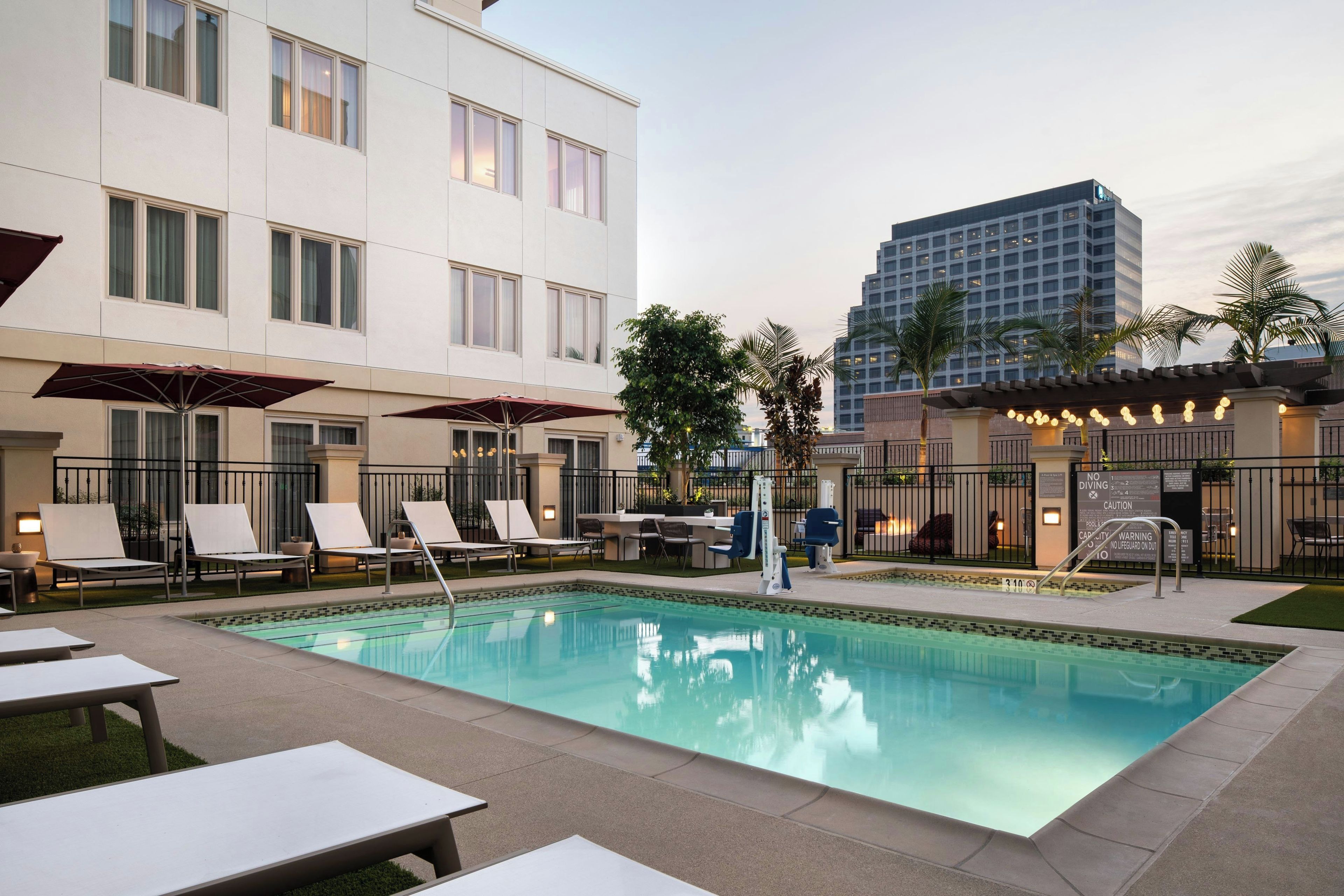 A swimming pool with lounge chairs and umbrellas, surrounded by a building with multiple windows and a tall structure in the background.