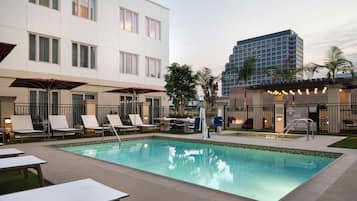 A swimming pool with lounge chairs and umbrellas, surrounded by a building with multiple windows and a tall structure in the background.