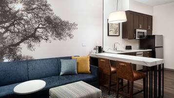 A modern living room with a blue sofa, a small round table, and a striped ottoman. A kitchen area with dark cabinets, a microwave, and a refrigerator is visible in the background.