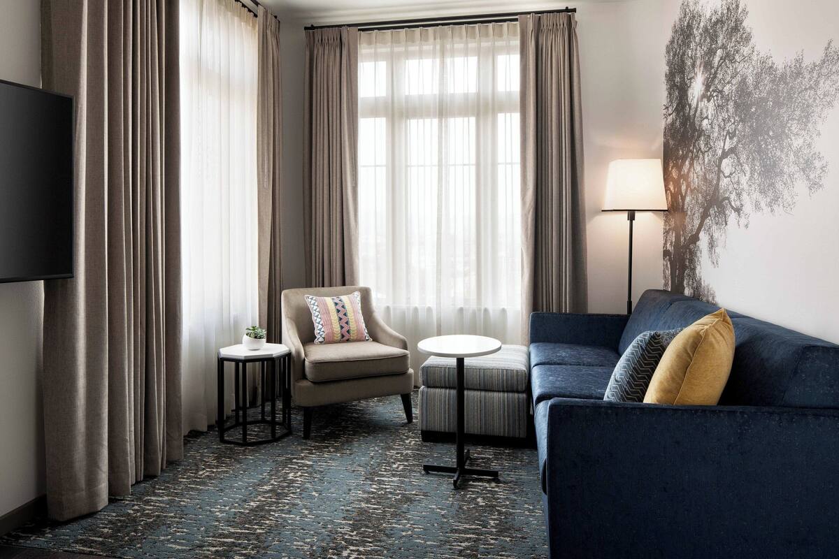 Habitación de hotel con sofá azul oscuro, una mesita redonda pequeña, un sillón color beige, televisor y un ventanal con cortinas traslúcidas.