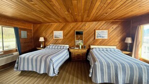 A cabin room with two beds, wooden walls, a wooden ceiling, a window with curtains, and a small table with a lamp.