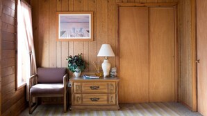 A wooden room with a chair, a small table with a lamp, a framed picture on the wall, and a door.