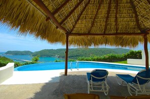 Un espace piscine doté d’un toit de chaume, de deux chaises longues et offrant une vue sur la mer et les collines.