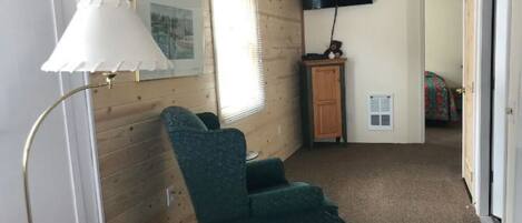 A room with a teal armchair, a white lamp, a wooden cabinet, and a television on the wall.