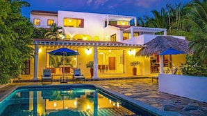 Una casa moderna de dos pisos con piscina, mobiliario exterior y estructura de techo de paja.