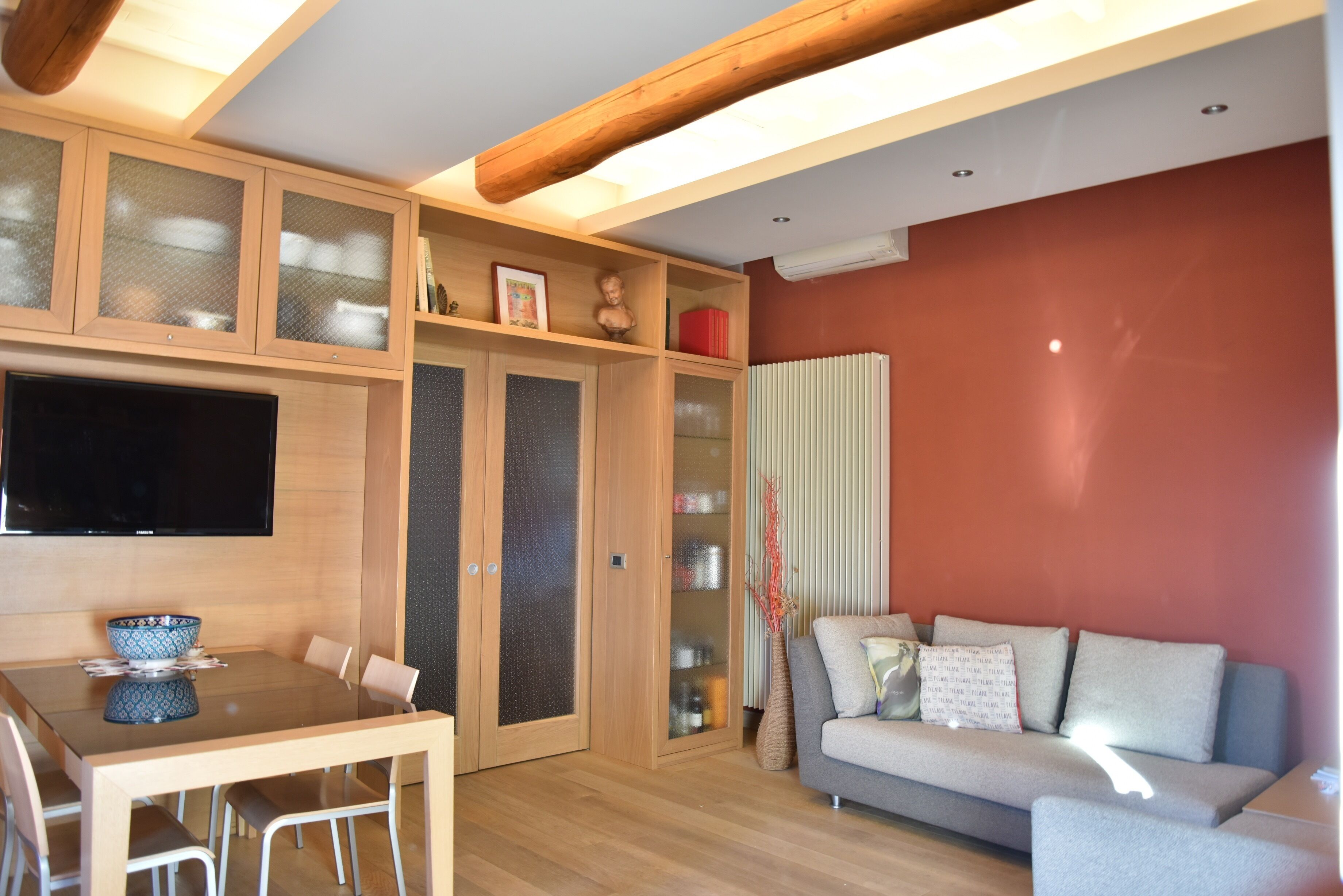 A modern living room with a wooden dining table, a grey sofa, and a TV mounted on the wall.