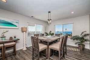 A dining area with a wooden table and chairs, a view of the ocean through large windows, and a ceiling light fixture.