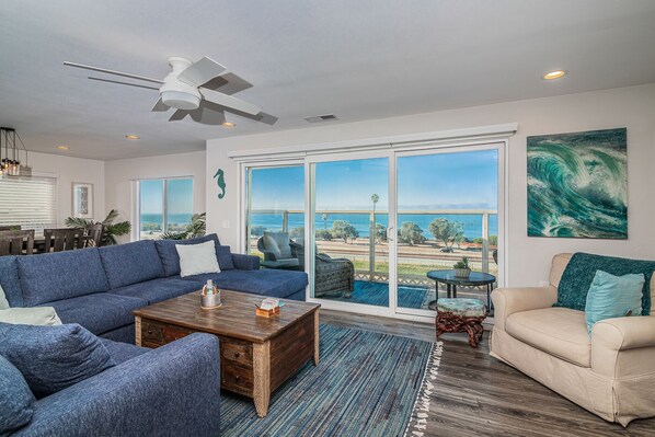 A spacious living room with a blue sofa, beige armchair, and a wooden coffee table. A ceiling fan and large windows provide a view of the ocean.