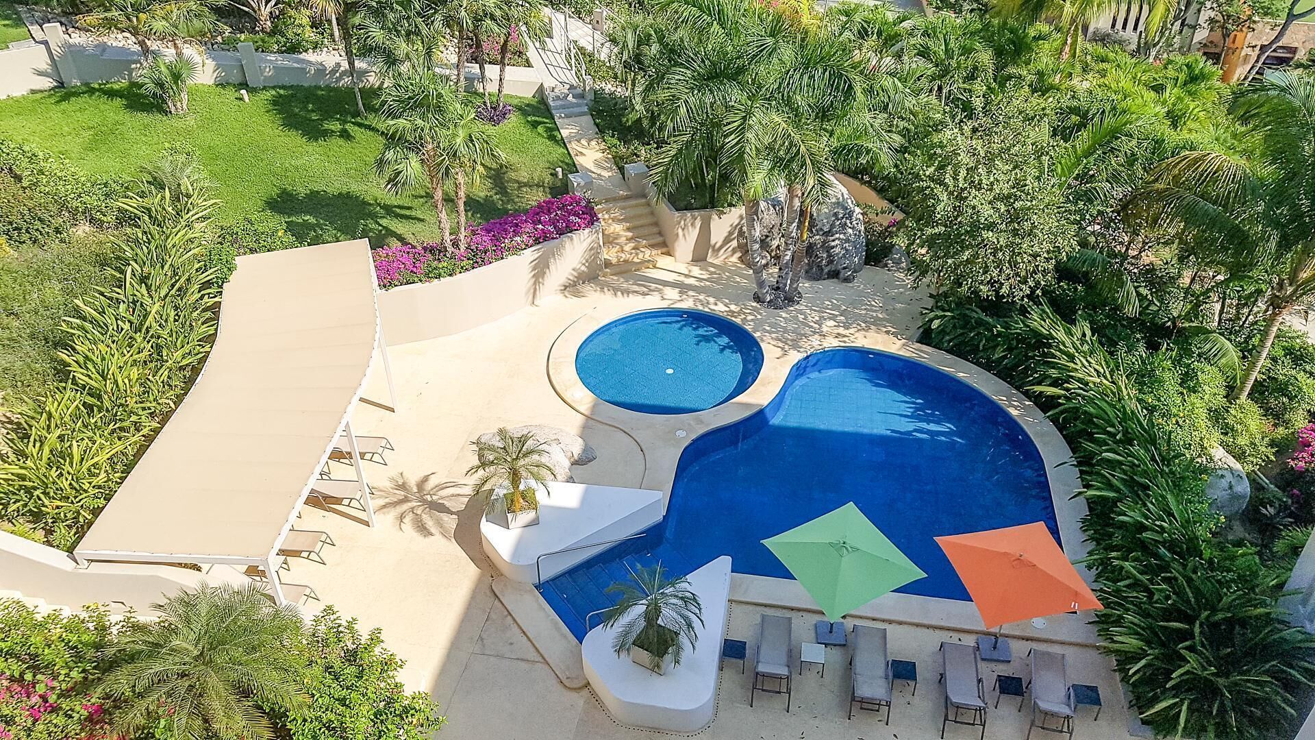 Une piscine dotée d’une partie plus profonde, entourée de chaises longues et de parasols, avec des palmiers et un jardin en arrière-plan.
