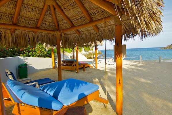 A beachside lounge with thatched roof, two blue cushioned lounge chairs, and a view of the sea.
