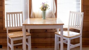 A wooden cabin interior with a table and two chairs, a window with curtains, and a vase with flowers on the table.