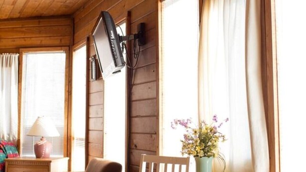 A wooden cabin interior with a dining table, chairs, a lamp, and a vase with flowers.