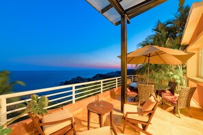 Uma varanda com vista para o oceano, equipada com cadeiras e uma mesa, e um grande guarda-sol.