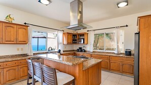 A spacious kitchen with wooden cabinets, a central island with bar stools, a stainless steel range hood, and a view of the sea through large windows. - Villa Diamante (Santa María Huatulco)