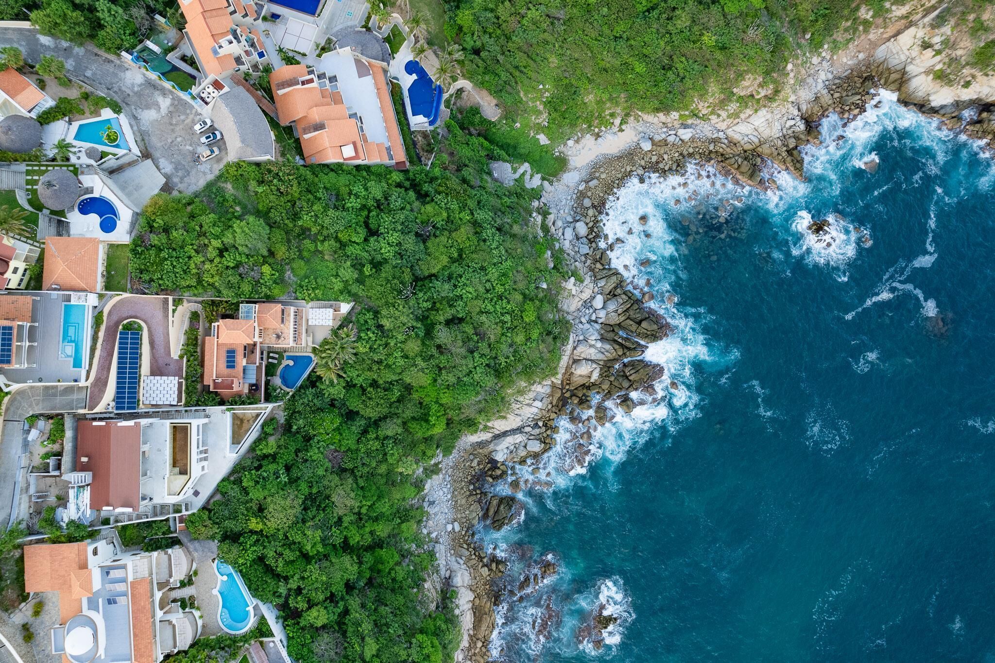 An aerial view of a coastal property with multiple swimming pools, lush greenery, and rocky shoreline.