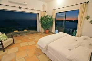 Un dormitorio con una cama grande, un balcón con vista a la ciudad al atardecer y una planta en maceta.