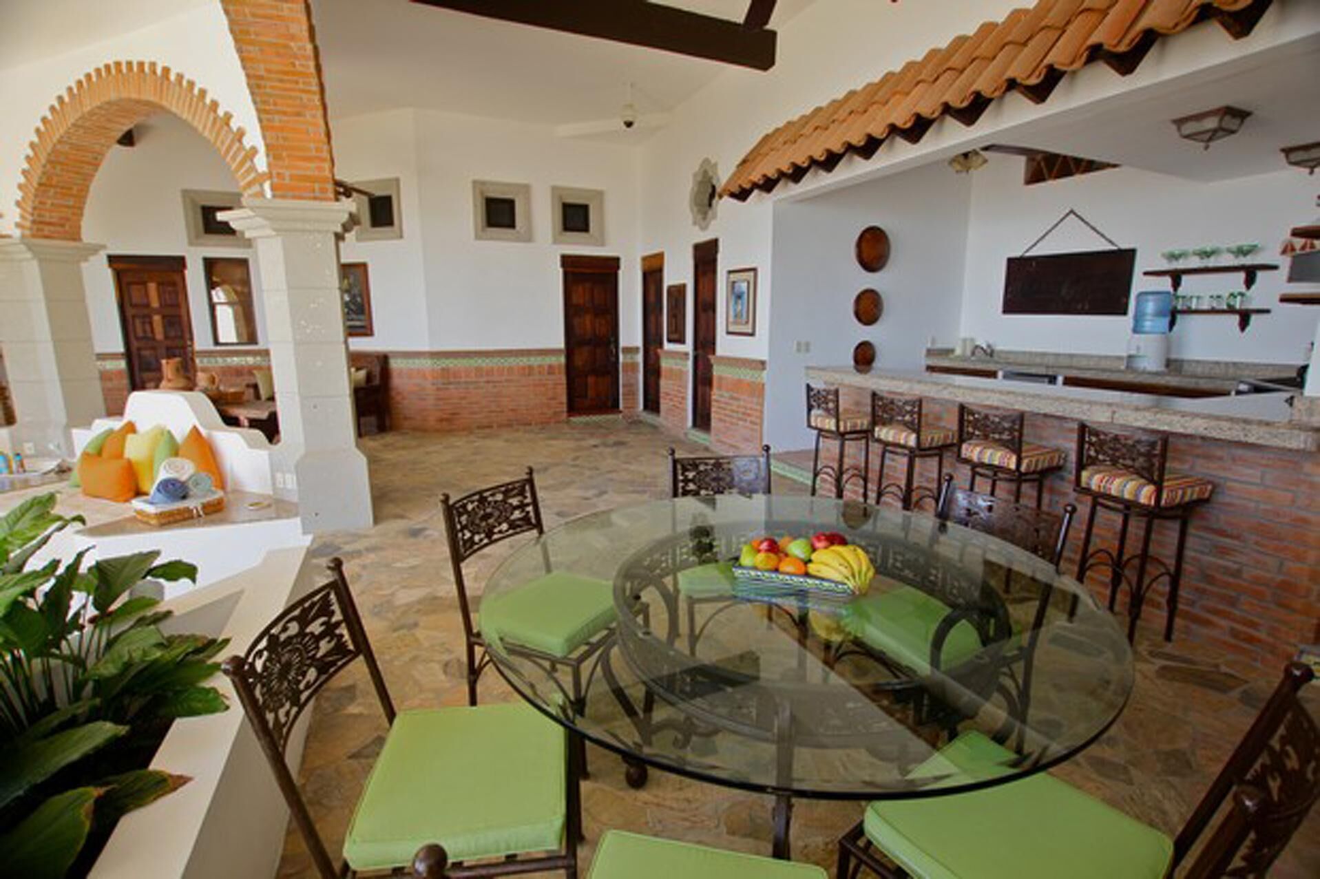 Un comedor amplio con una mesa de vidrio rodeada de sillas, una zona de cocina con barra y un patio con pared y arco de ladrillo.