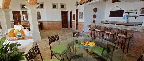 Un comedor amplio con una mesa de vidrio rodeada de sillas, una zona de cocina con barra y un patio con pared y arco de ladrillo.