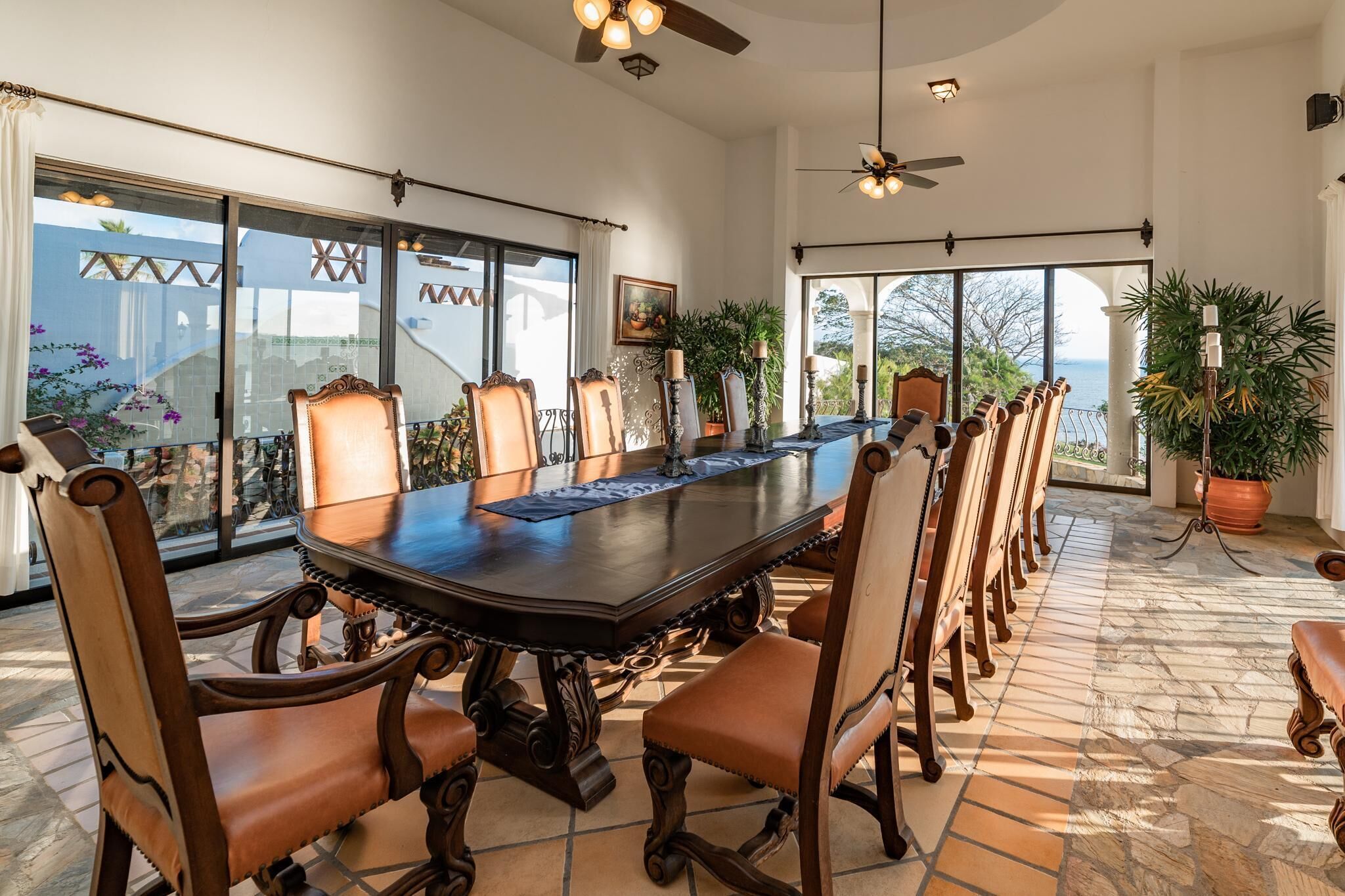 A large dining table with ornate chairs, large windows, and ceiling fans in a spacious room.