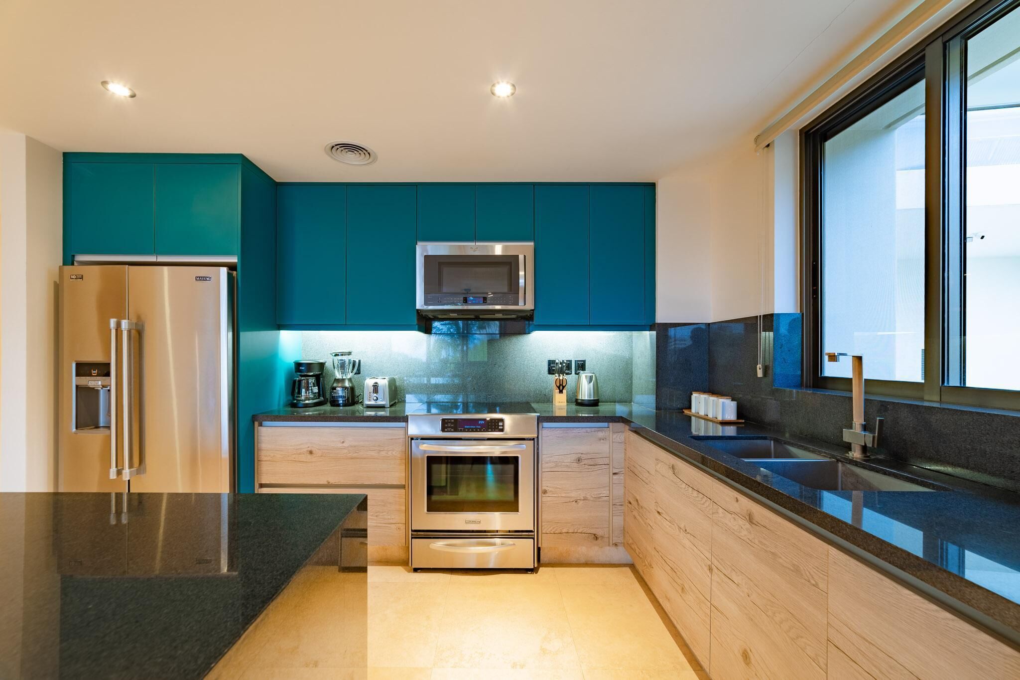 A modern kitchen with teal cabinets, stainless steel appliances, and a large window providing natural light.