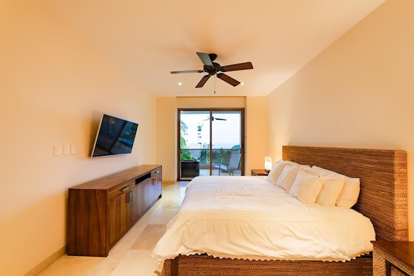 A bedroom with a bed, a TV on the wall, a ceiling fan, and a view through a sliding door.