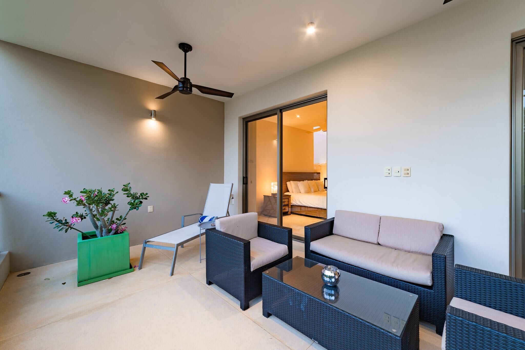 A modern outdoor seating area with wicker furniture, a ceiling fan, and a green planter with flowers.