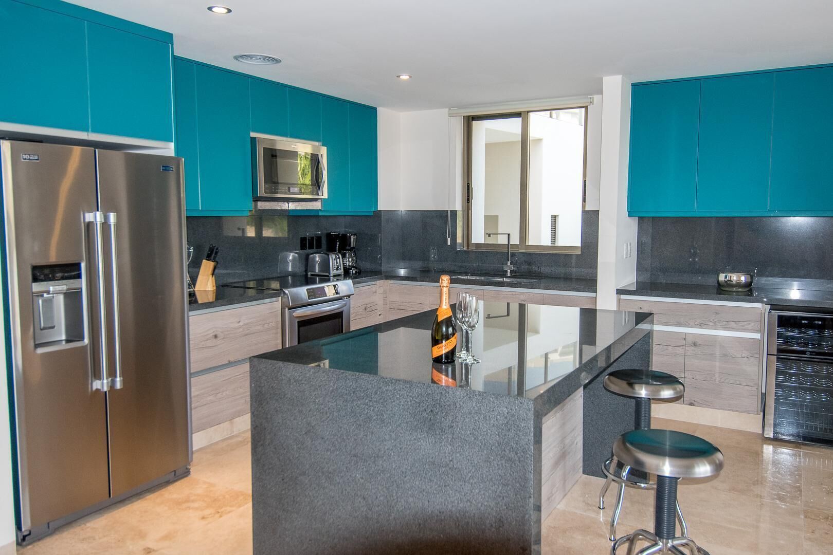 A modern kitchen with teal cabinets, stainless steel appliances, a central island with stools, and a large window.