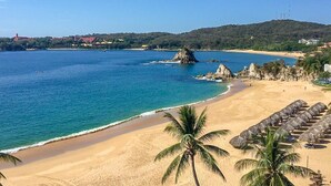 Une plage à l’eau d’un bleu limpide, avec un rivage sablonneux, des palmiers et des parasols en feuilles de palmier.