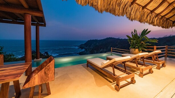 An infinity pool with wooden lounge chairs and a table, overlooking the ocean at dusk. - Villa Alheli (Santa María Huatulco)