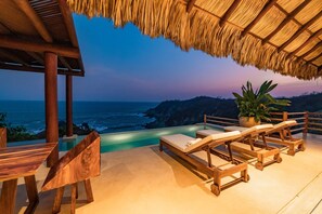 An infinity pool with wooden lounge chairs and a table, overlooking the ocean at dusk. - Villa Alheli (Santa María Huatulco)