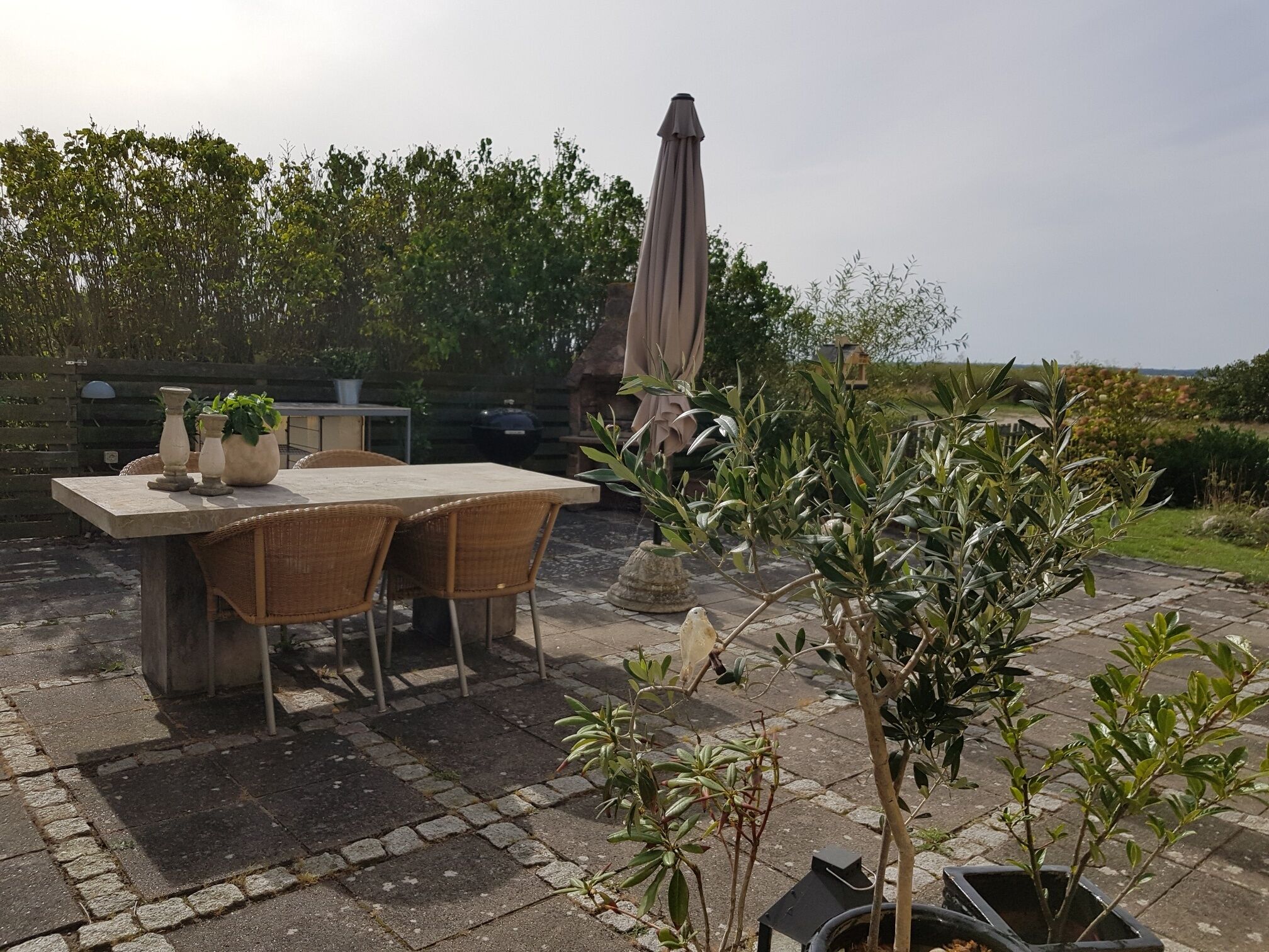 An outdoor patio with a stone table and wicker chairs, surrounded by potted plants and a large umbrella.