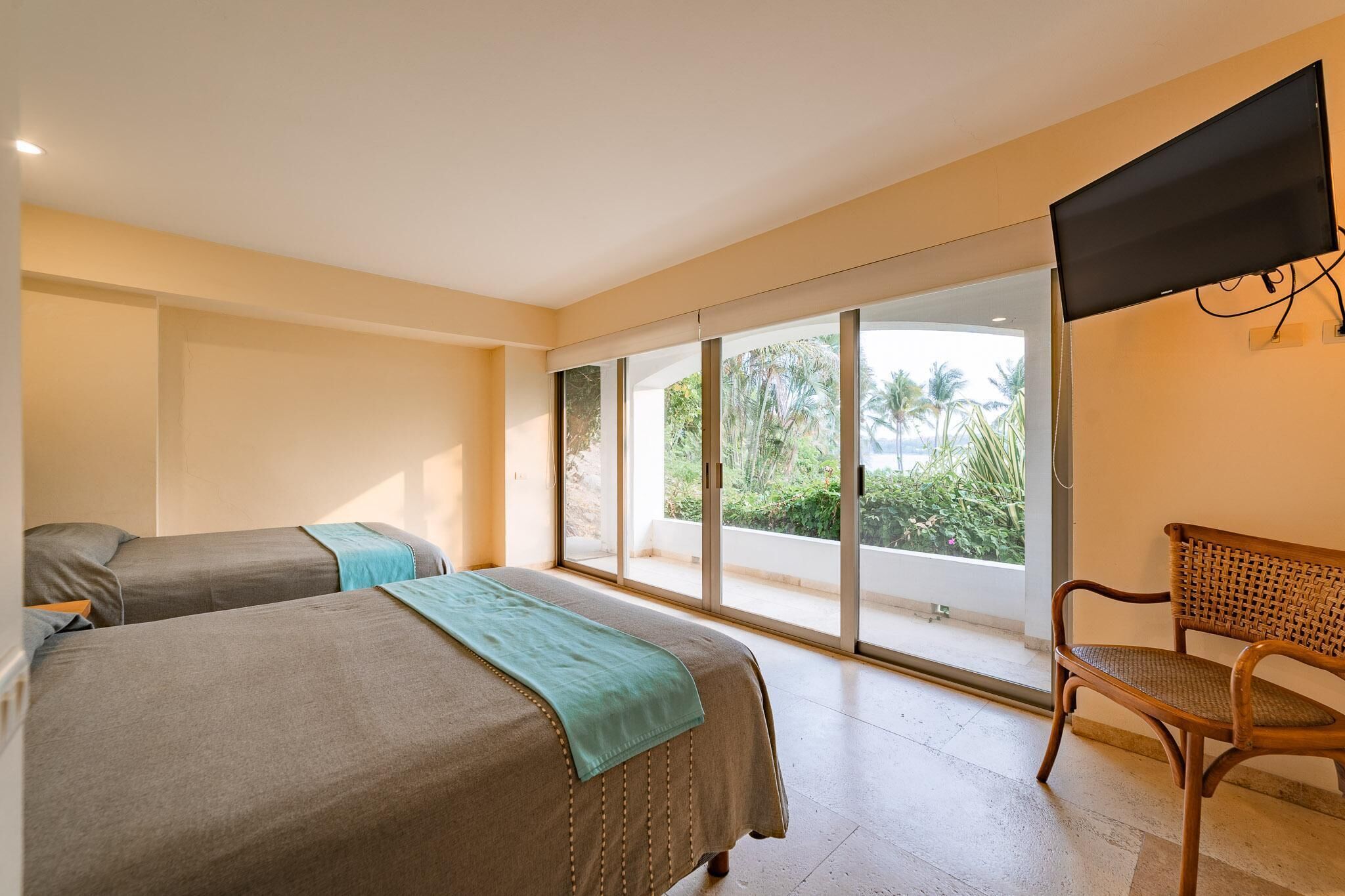 Habitación de hotel con dos camas, un televisor de pantalla plana, una silla de mimbre y un amplio ventanal corredero que da a un balcón con vista a palmeras.