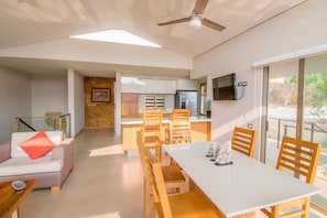 A modern living room with a sofa, a dining table, and chairs. There is a kitchen area with cabinets and a refrigerator. A ceiling fan is visible, and there is a TV mounted on the wall.